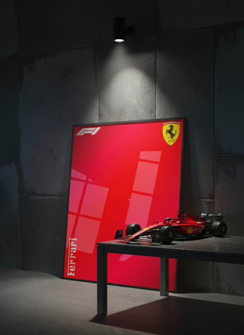 A large vertical red Ferrari poster or print leaning against a dark gray wall, with a small red Ferrari Formula 1 car placed on a table in front of it. A spotlight shines on the wall above the setup.